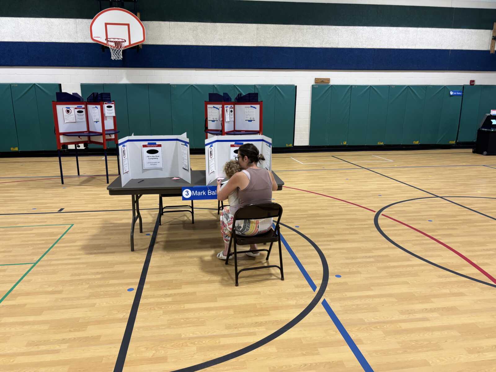 Polling station set up in a gym with privacy booths and a voter sitting with a child at a table labeled '3' to vote or fill out forms.