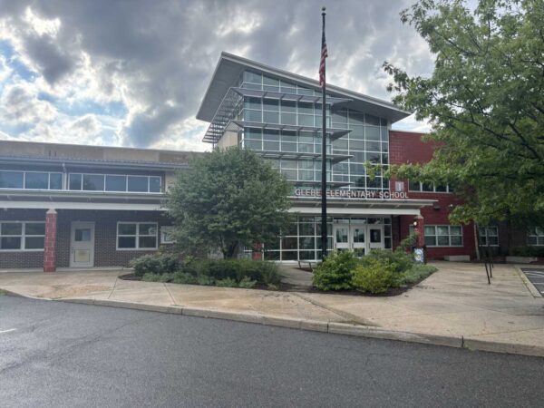 Front view of an elementary school with a tall flagpole, glass atrium, and landscaped bushes outside the entrance.