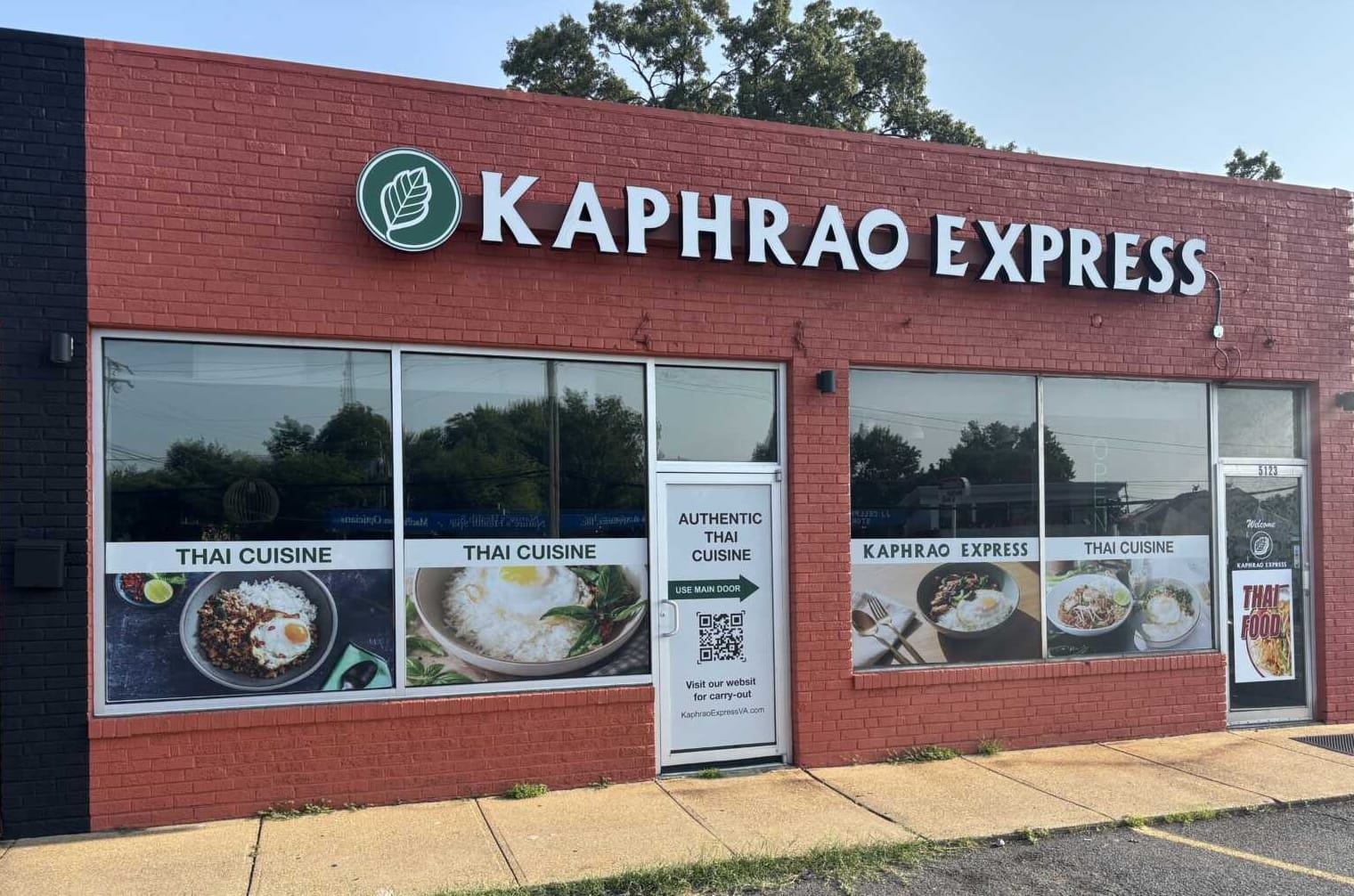 Red brick storefront with large white 'KAPHRAO EXPRESS' sign and a green circular leaf logo, windows showing Thai cuisine banners outside.