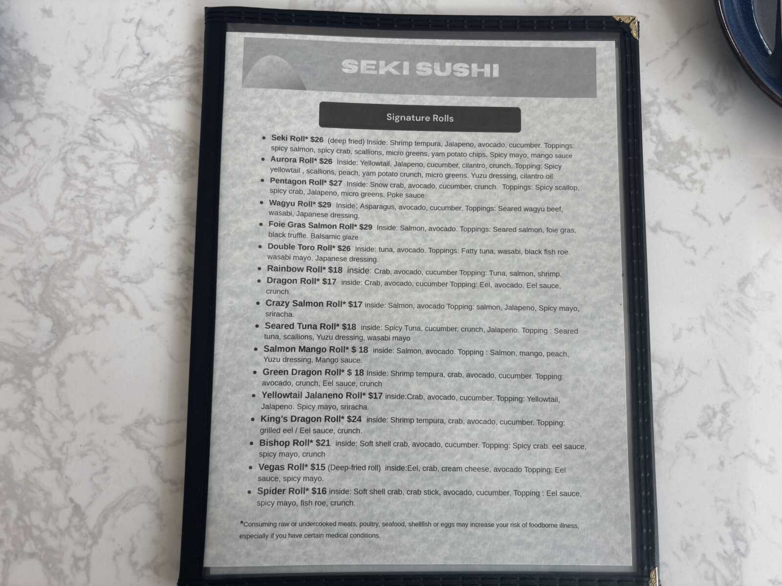 Menu page in a black folder on a marble table, showing 'SEKI SUSHI' header and a 'Signature Rolls' list of sushi options.