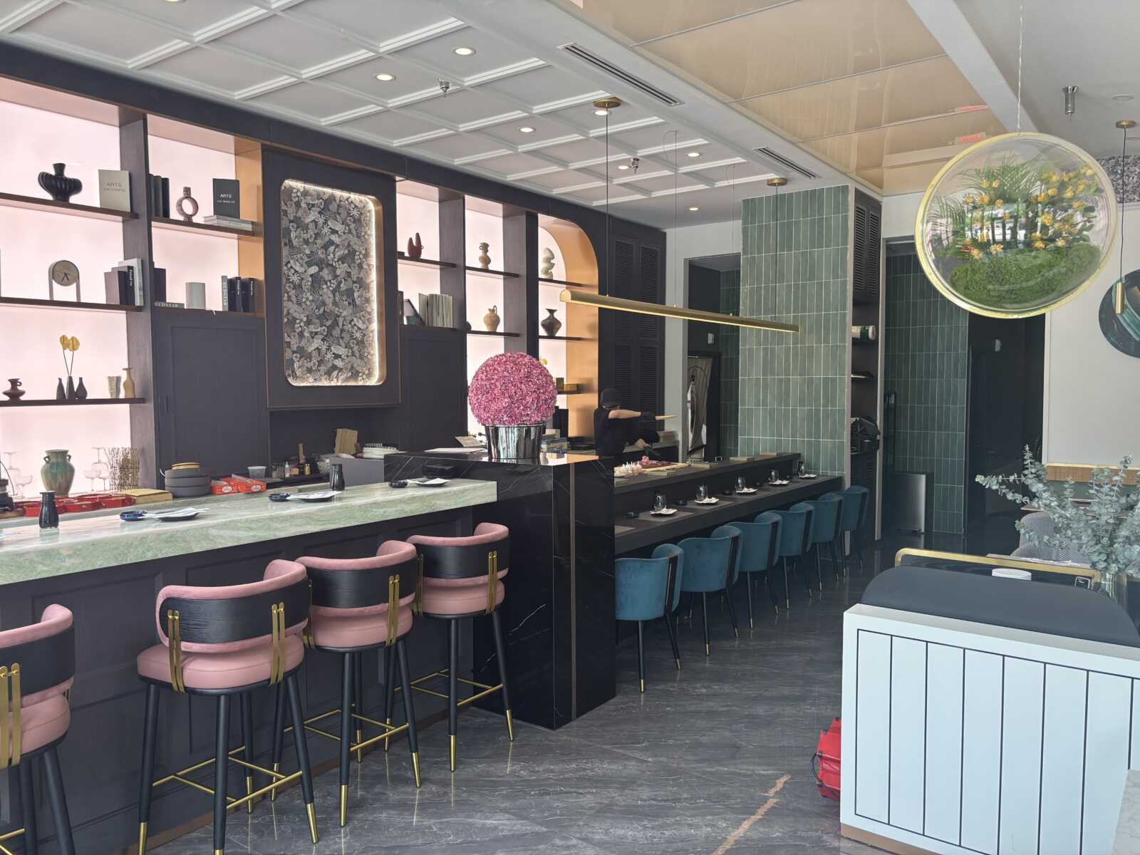 Modern cafe interior with a long marble counter, pink and teal bar stools, and decorative shelves along the wall.