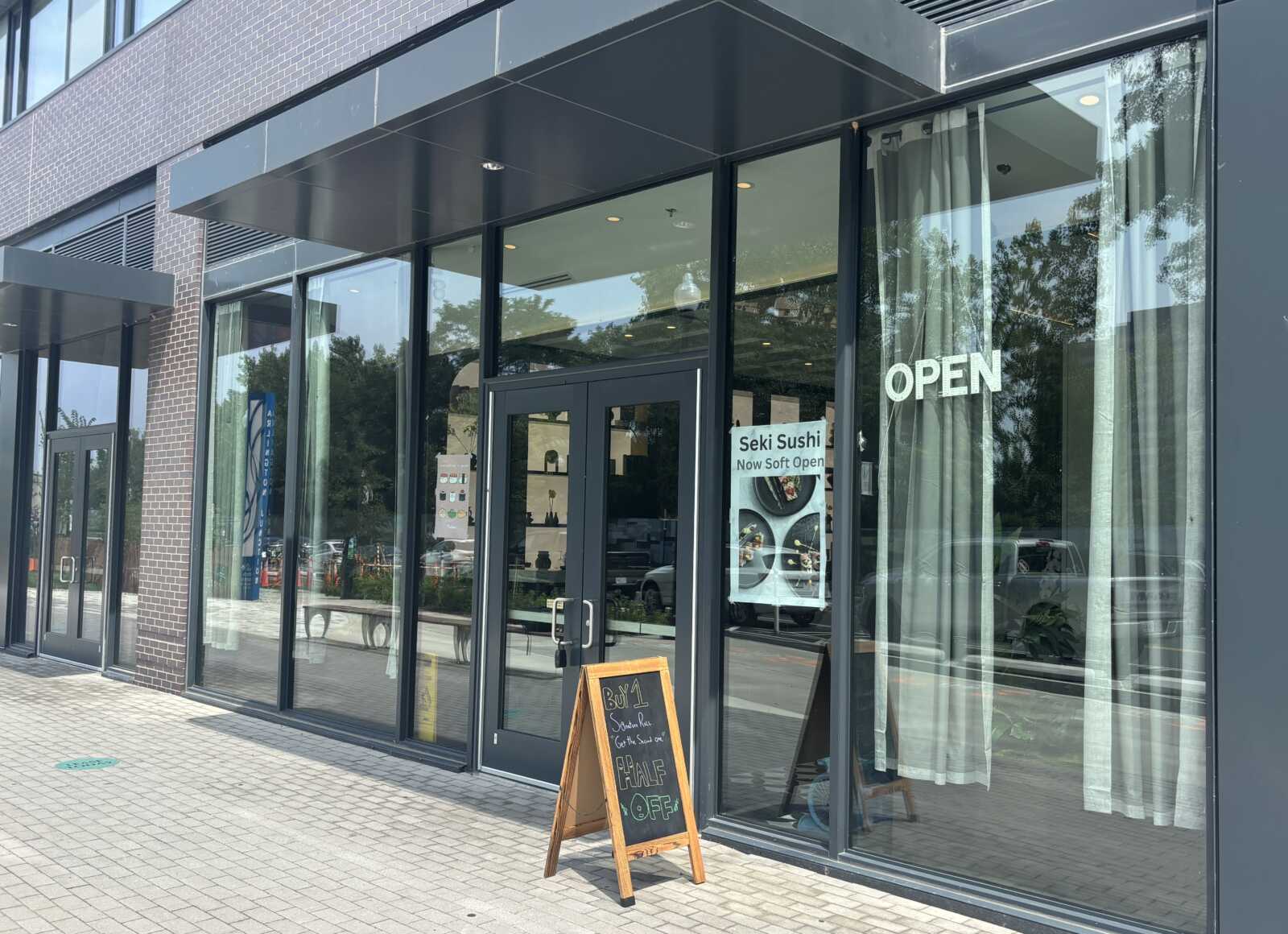 Storefront of a sushi restaurant with large glass doors, an 'OPEN' sign in the window, and a chalkboard promo sign outside.