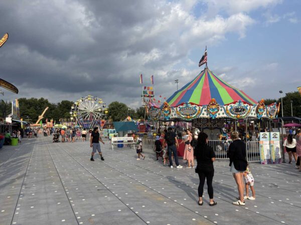 Colorful carnival scene with a striped big-top tent, a Ferris wheel, and visitors strolling along a paved midway on a sunny day.