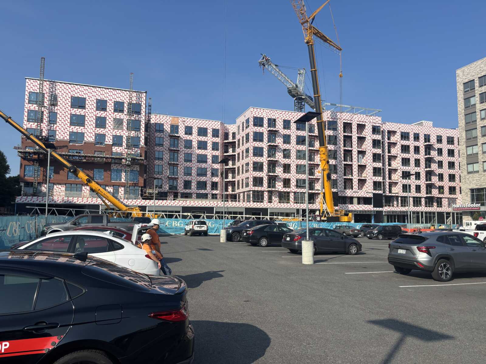 Construction site with a multi-story building under cladding, two yellow cranes overhead, and a parking lot in the foreground.