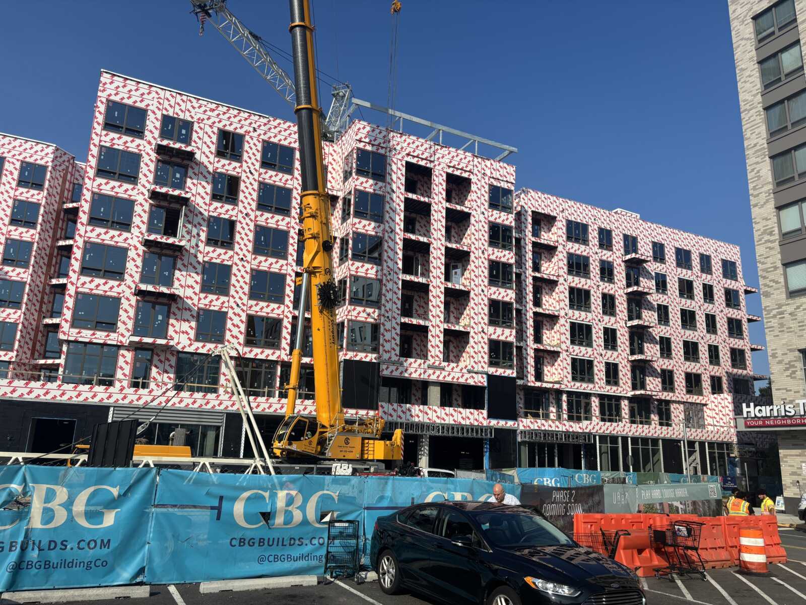 Construction site with a multi-story building wrapped in white weatherproofing with red logos, and a large yellow crane in the foreground under a clear blue sky.