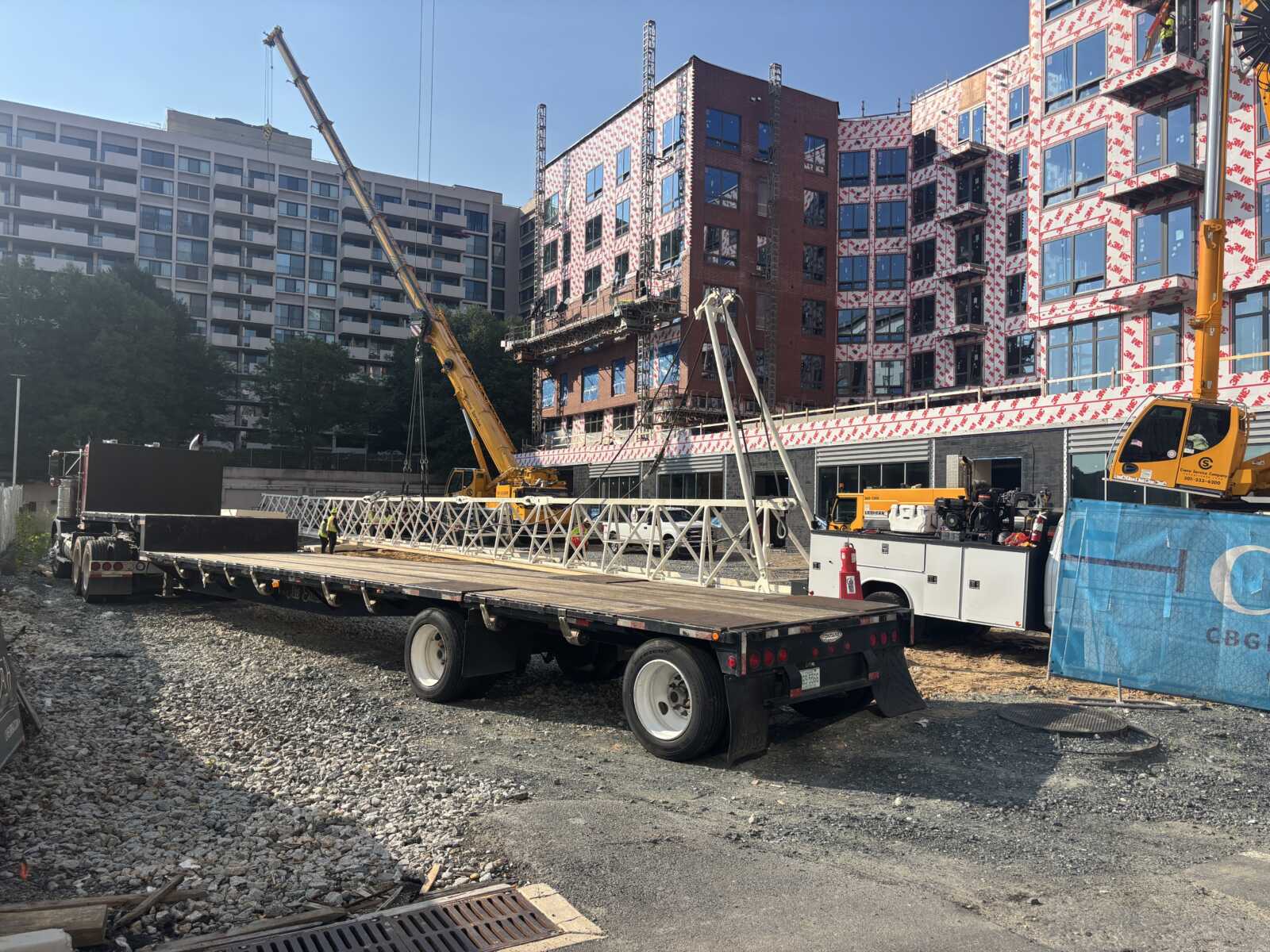 Construction site with a large flatbed truck and crane lifting a metal framework near a multi-story building under construction.