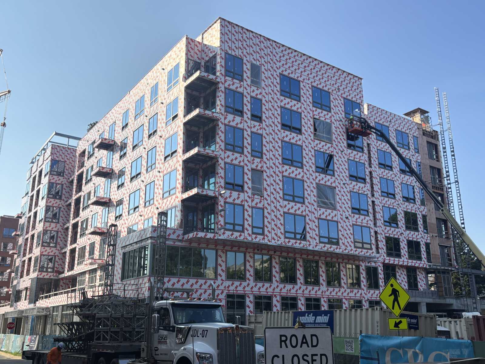 Under-construction, multi-story building wrapped in protective material with scaffolding and construction equipment on site, blue sky overhead.