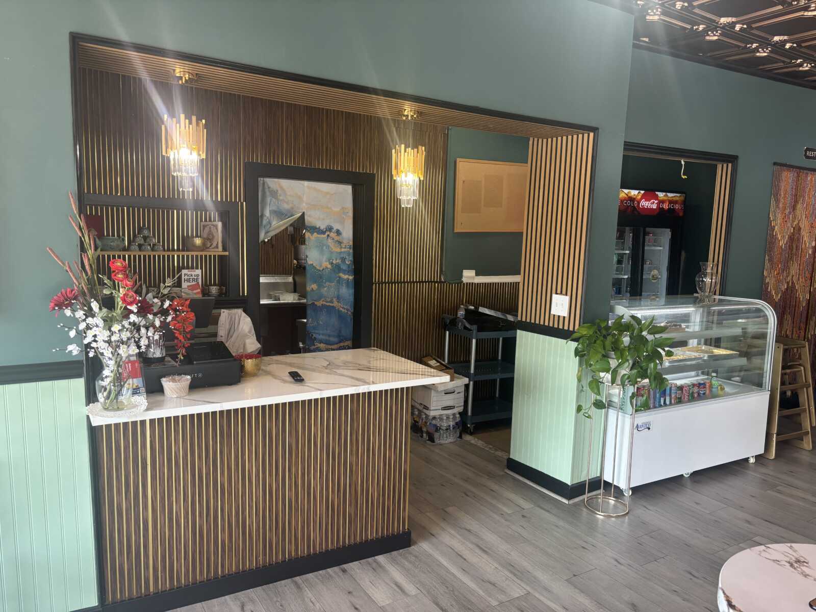 Cafe counter area with teal walls and gold striped panels, marble top, and a vase of red and white flowers beside a register.