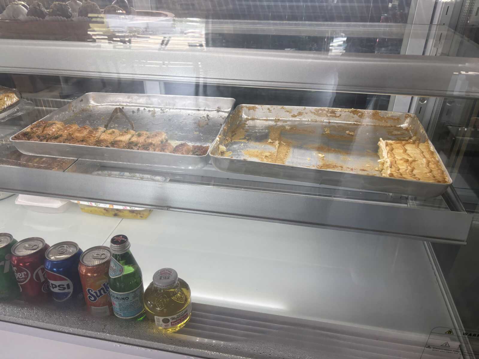 Metal display case with two large baking trays of partially eaten pastries; one tray mostly empty with scraps, the other with some cake pieces along the edge; reflections on glass.