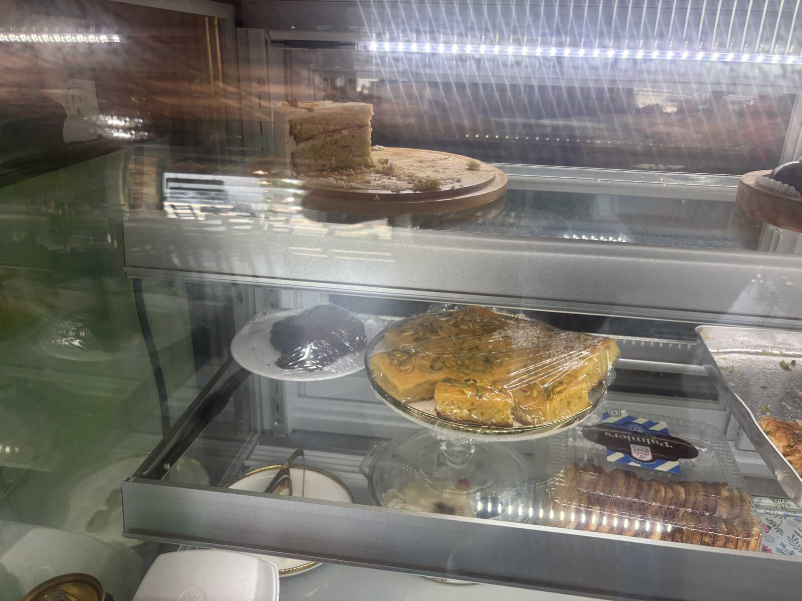 Cake with a slice removed on a round board in a glass bakery display case, reflections visible in the glass front