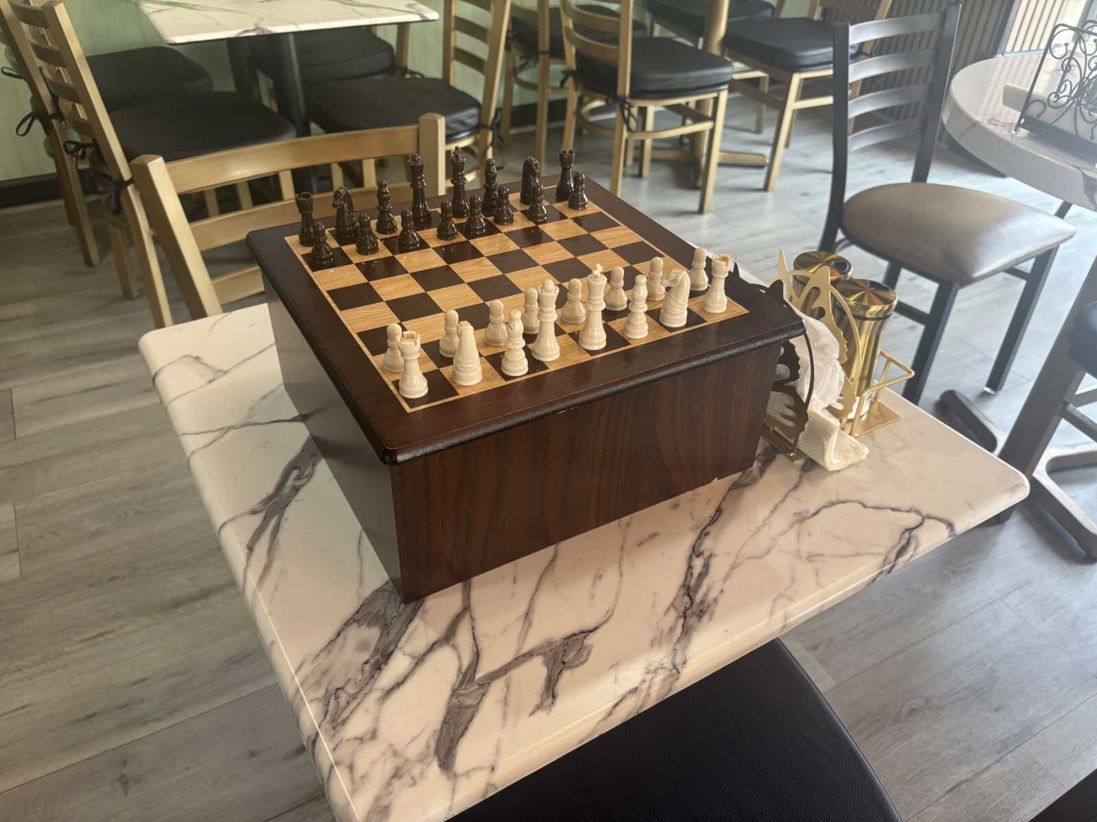 Chessboard with white and black chess pieces arranged for a match on a marble-top table in a cafe, with chairs in the background and trophies nearby.