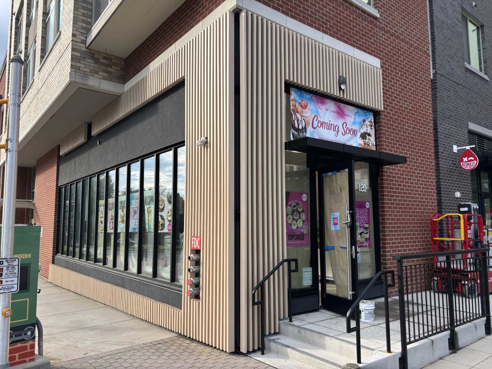 Storefront with large glass windows and a beige vertical-slat facade on the corner of a red brick building; a 'Coming Soon' banner above the entrance.