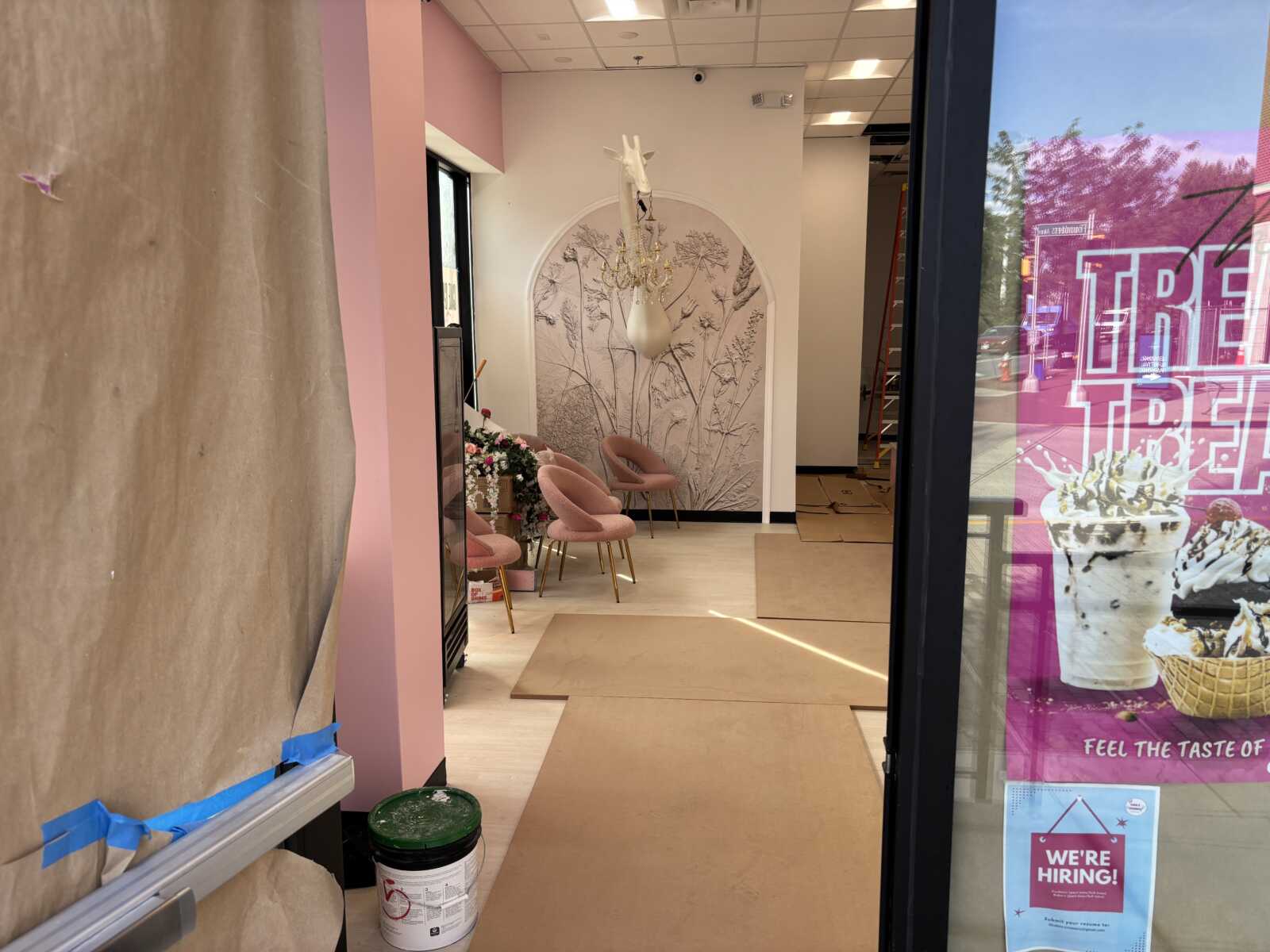 Store interior under renovation with pink walls, a white botanical mural, pink chairs, and a hanging chandelier, plus construction materials on the floor.