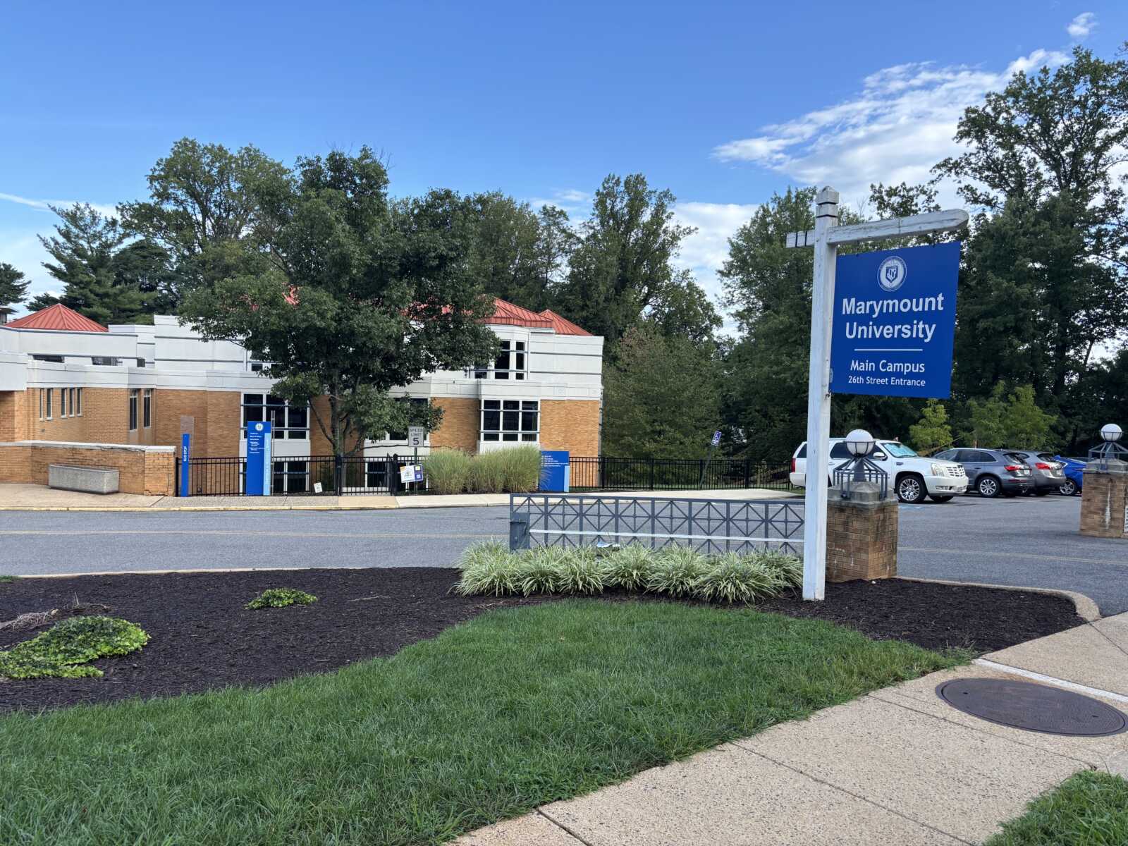 Blue Marymount University sign reading 'Main Campus' near the 26th Street Entrance by a parking lot and a modern brick building behind it