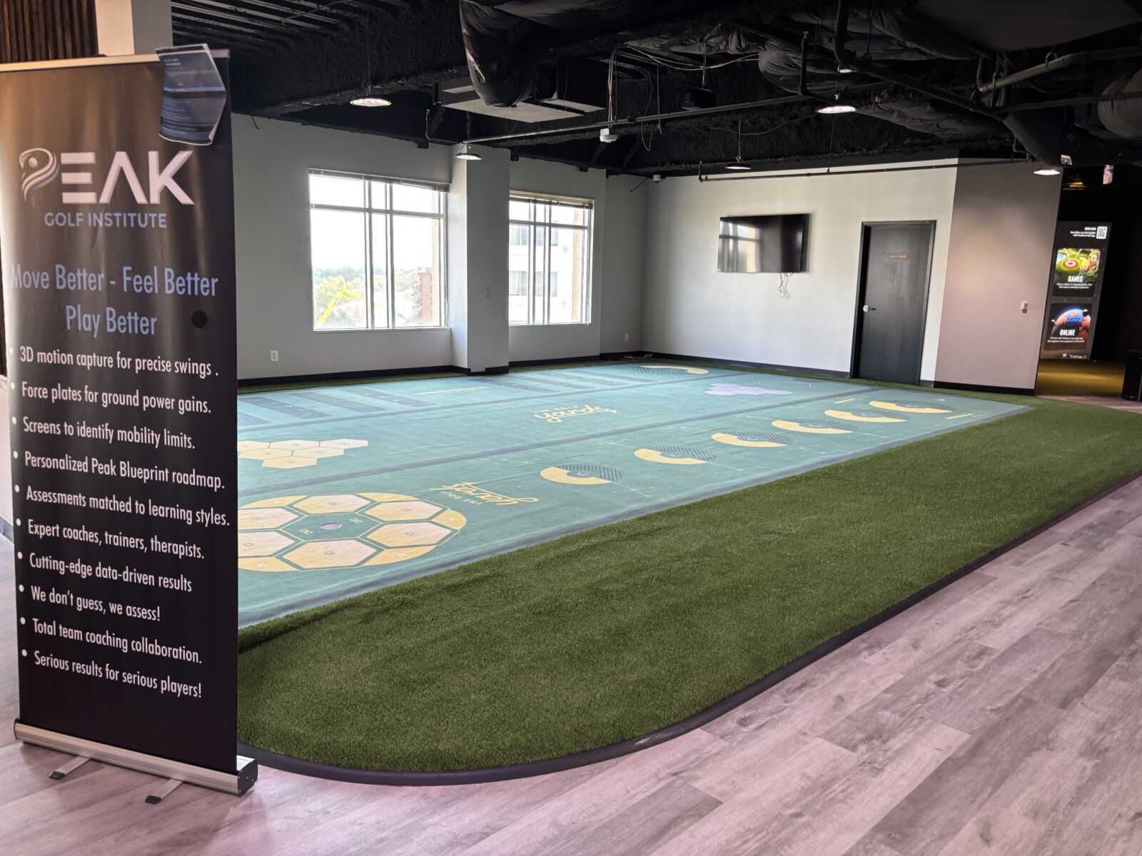 Indoor golf training area with large green turf, hexagonal patterns, and a tall banner reading 'PEAK GOLF INSTITUTE' on the left.