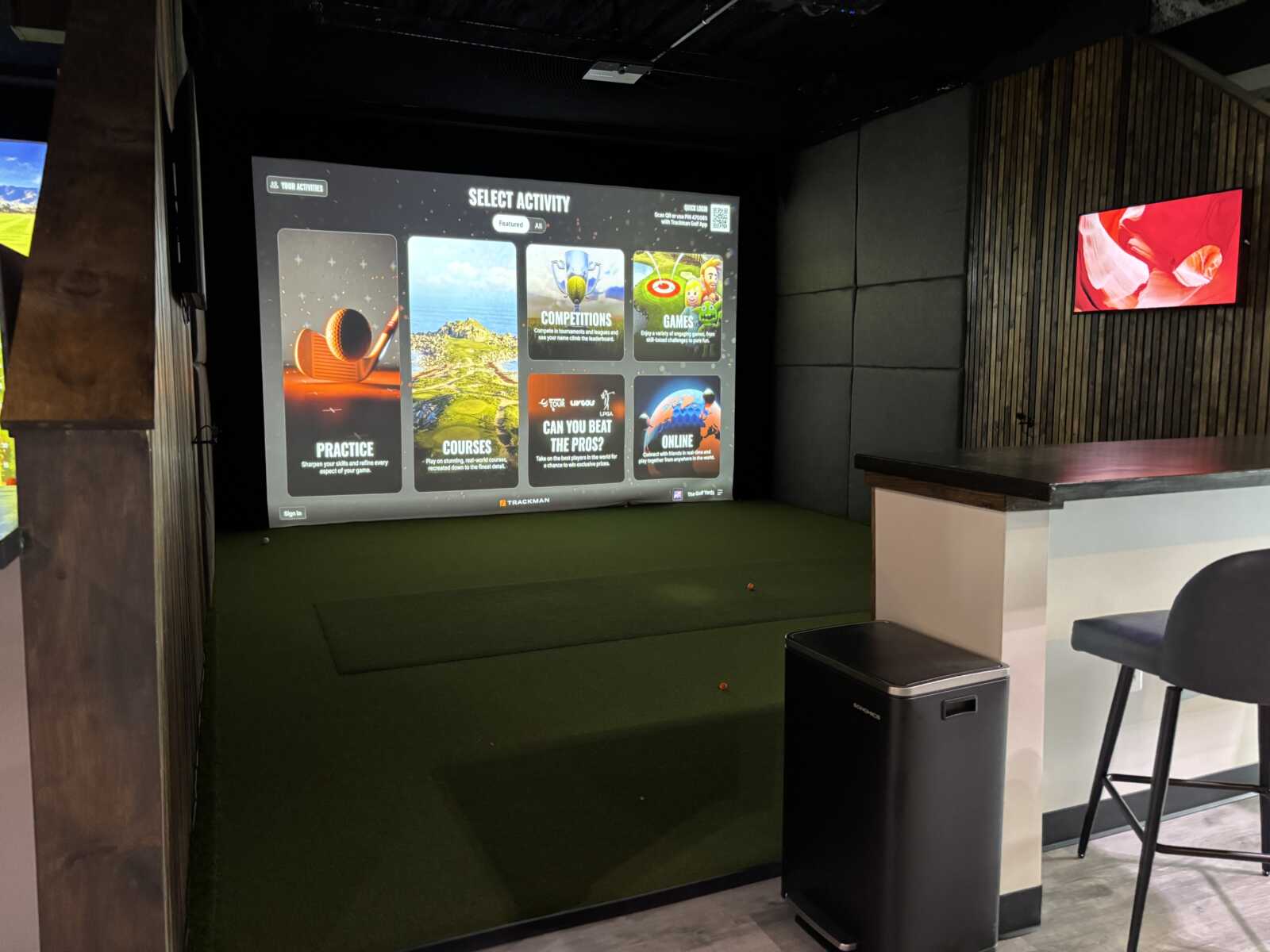 Indoor golf simulator with a large screen menu titled 'Select Activity' showing options like Practice, Courses, Competitions, and Games in a dim indoor facility.