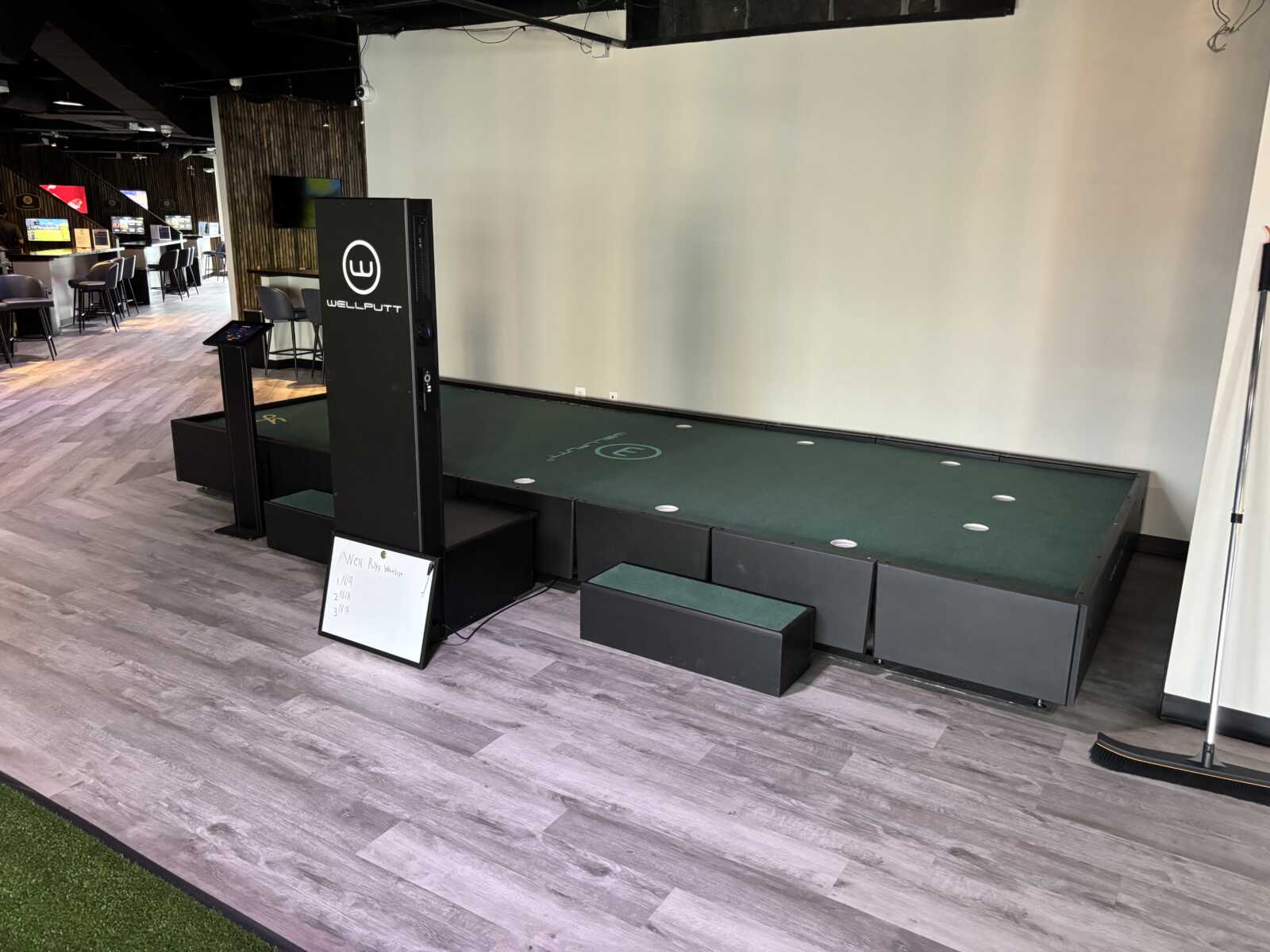 Indoor golf practice station with a green putting mat and a tall WellsPutt console in a lounge area