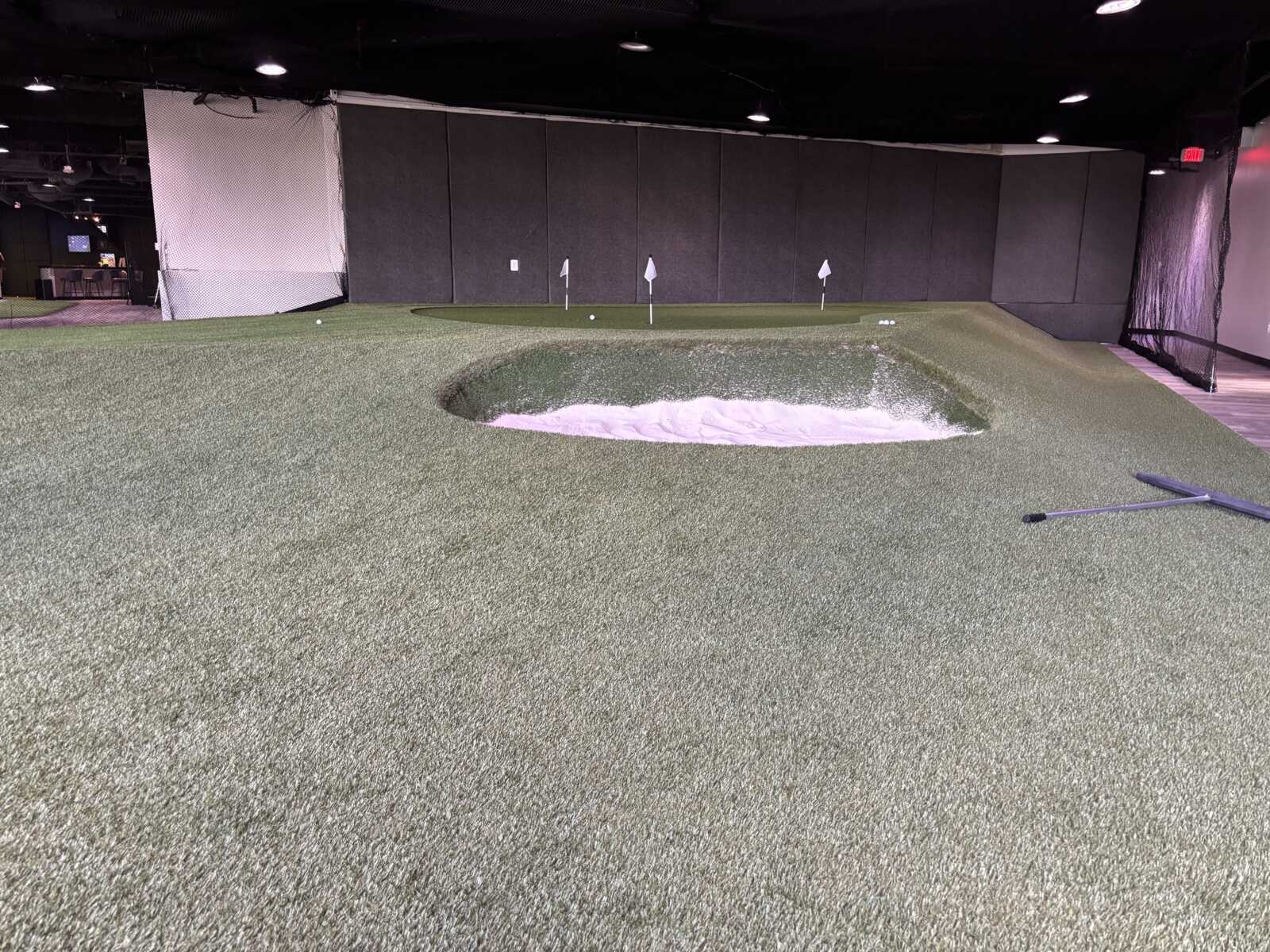 Indoor golf practice area with artificial turf, a sand bunker, and small flag markers near the green in a dim indoor facility.