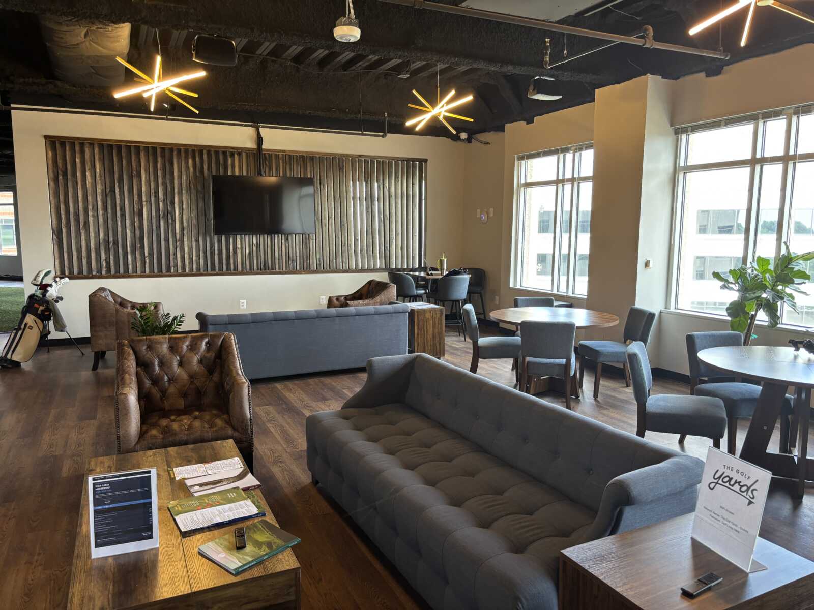 Modern lounge area with gray tufted sofa, brown leather chair, and round tables near large windows; TV mounted on a wooden slat wall to the left.