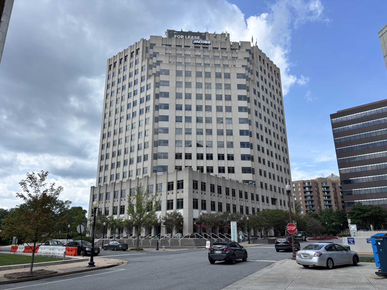 Tall beige office building with a 'For Lease Jacobs' sign on the roof, street in front with cars and orange construction barriers left.