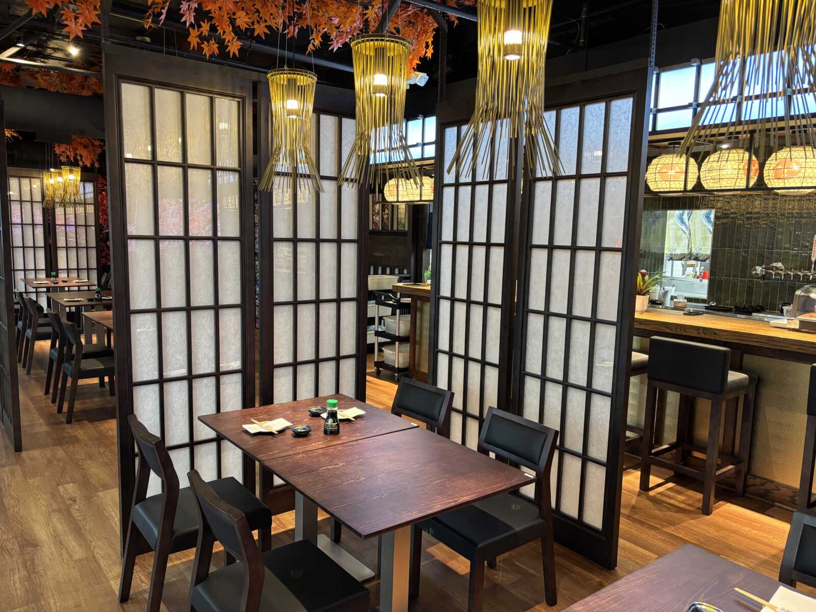 Restaurant interior with dark wood furniture, white frosted partition screens, and warm pendant lights; autumn leaves hang from the ceiling above.
