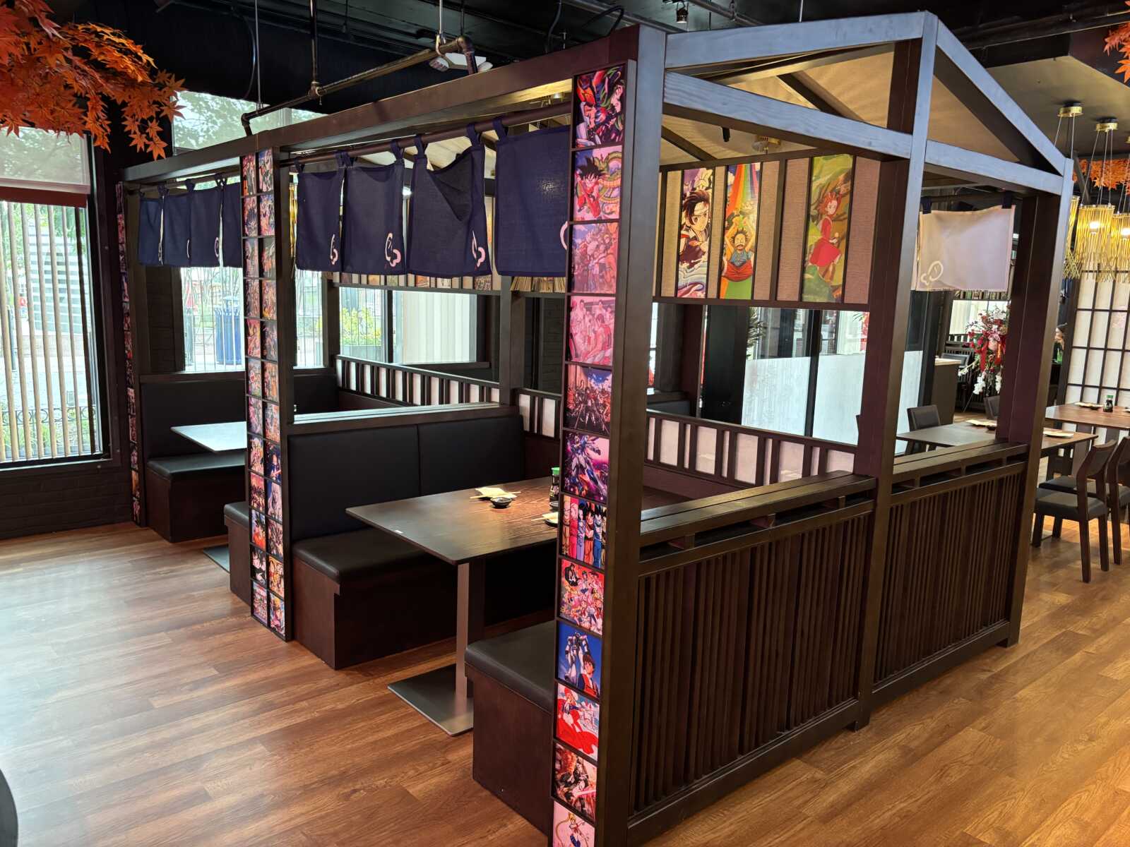 Dining booth area in a restaurant with dark wood partitions, black cushions, blue fabric dividers, and colorful anime panels along the sides.