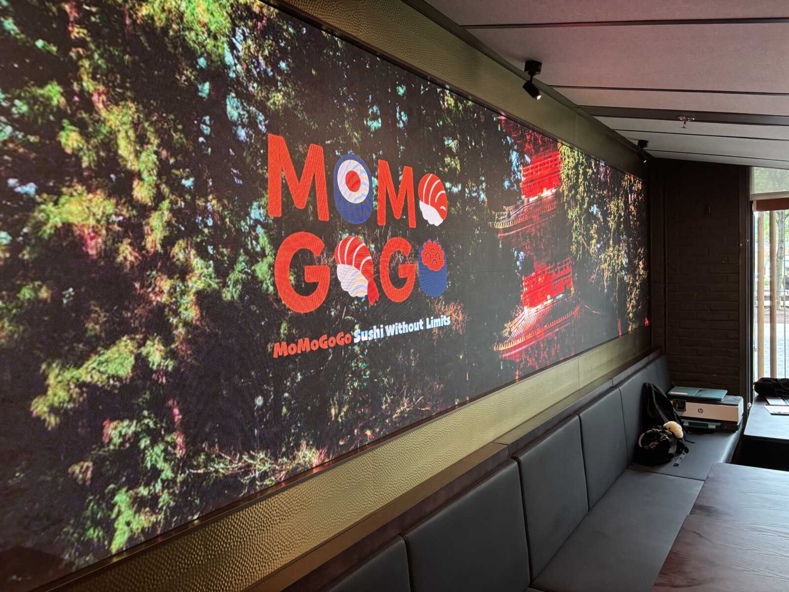Indoor seating area with a large digital wall display showing the MoMoGoGo sushi logo and 'Sushi Without Limits'.