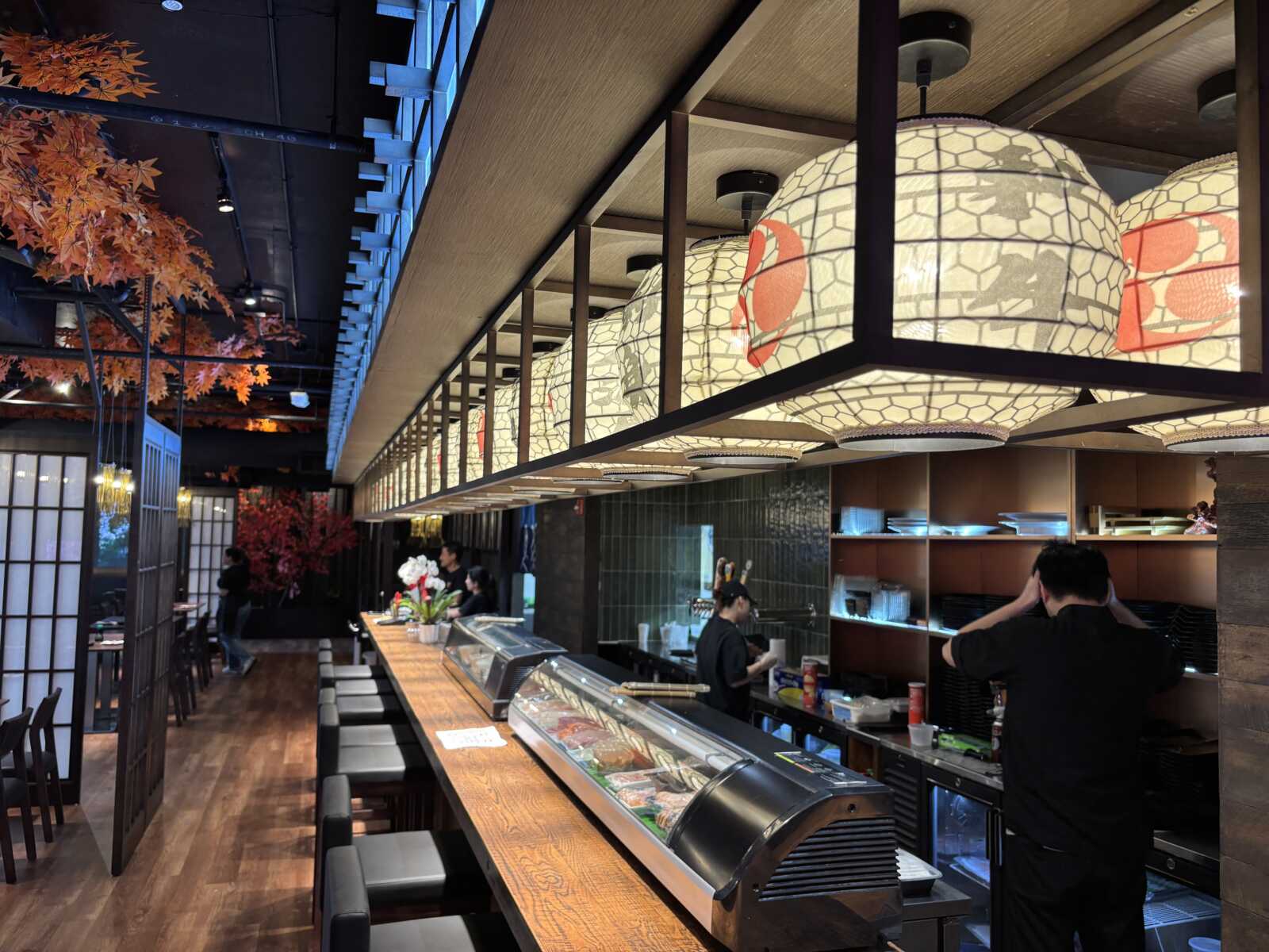 Interior of a modern sushi bar with a long wooden counter, stools, a glass sushi display, and warm hanging lanterns as staff prepare food.