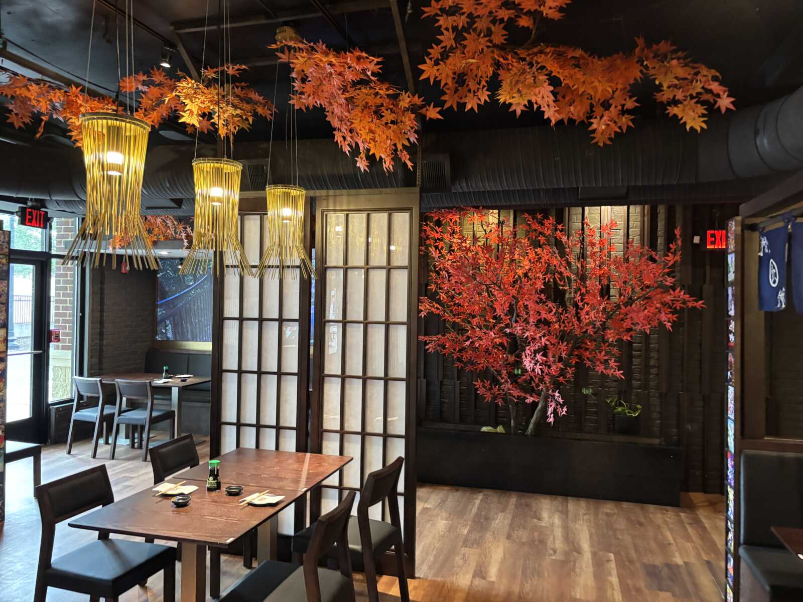 Restaurant interior with orange maple leaf decorations hanging from the ceiling, warm pendant lights, wooden screens, and a red-leaved tree centerpiece.