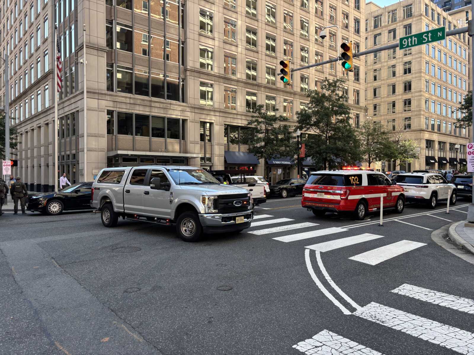 City intersection with crosswalk, tall office buildings, a gray pickup, a red emergency vehicle, and pedestrians nearby. 9th St N sign visible; urban street scene with traffic light.