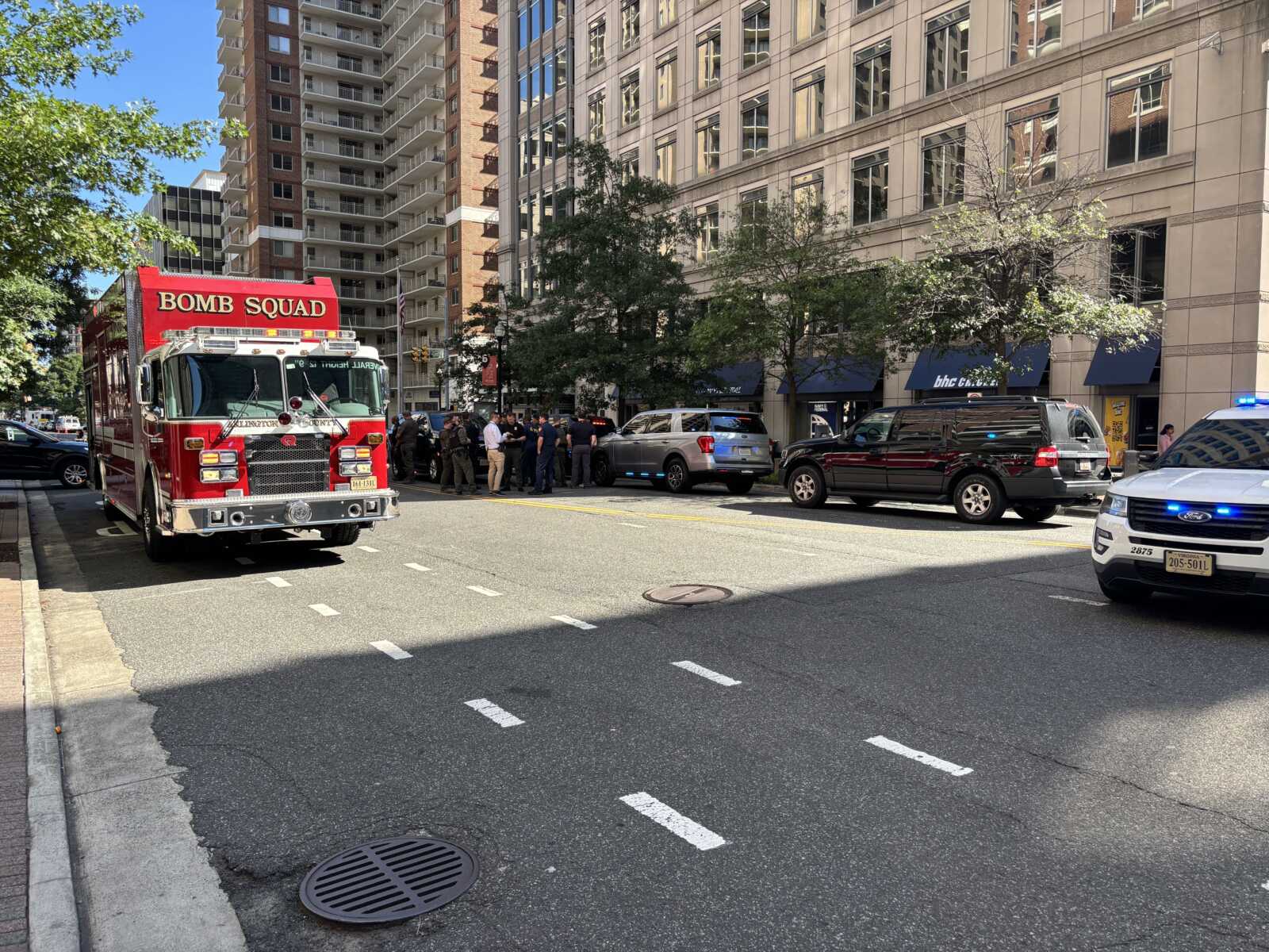 Red Bomb Squad fire truck parked on city street with a group of people nearby and tall buildings in the background.
