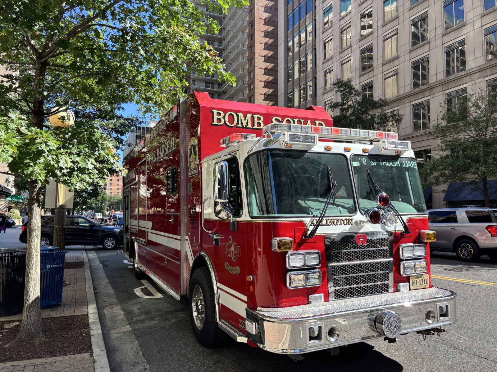 Red fire truck with chrome bumper parked on a tree-lined city street, tall buildings in the background.",
