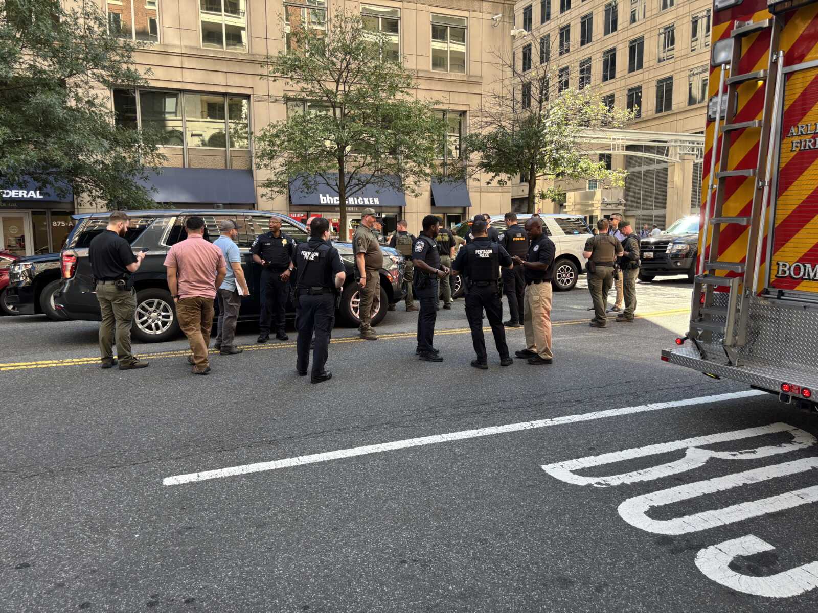 Group of police and emergency responders standing in a city street, discussing among parked vehicles and a fire truck nearby.
