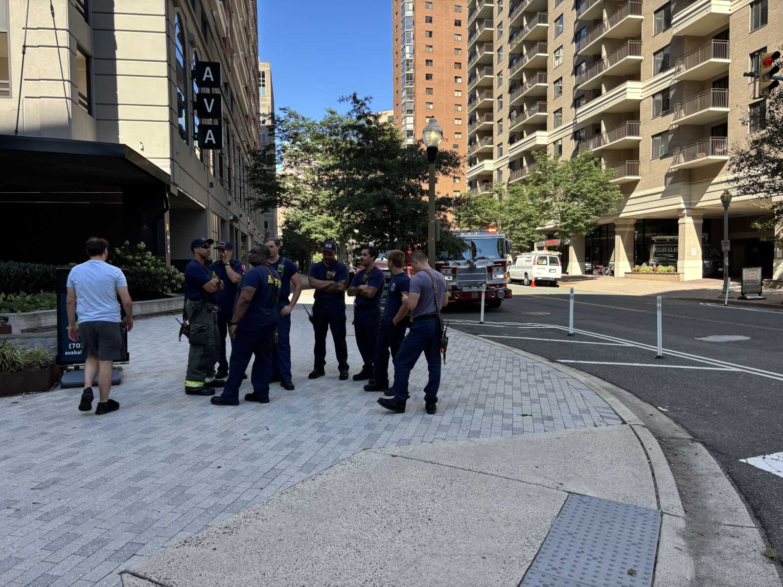 Firefighters chat on a city sidewalk with a fire engine and tall buildings in the background.