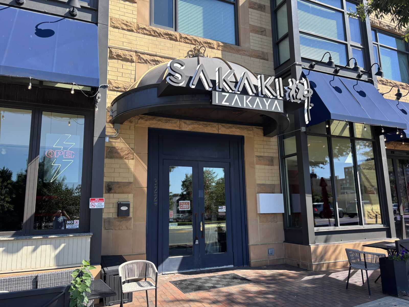 Front of a modern restaurant with a curved black awning and 'SAKAKI IZAKAYA' sign above double doors; outdoor seating to the left.