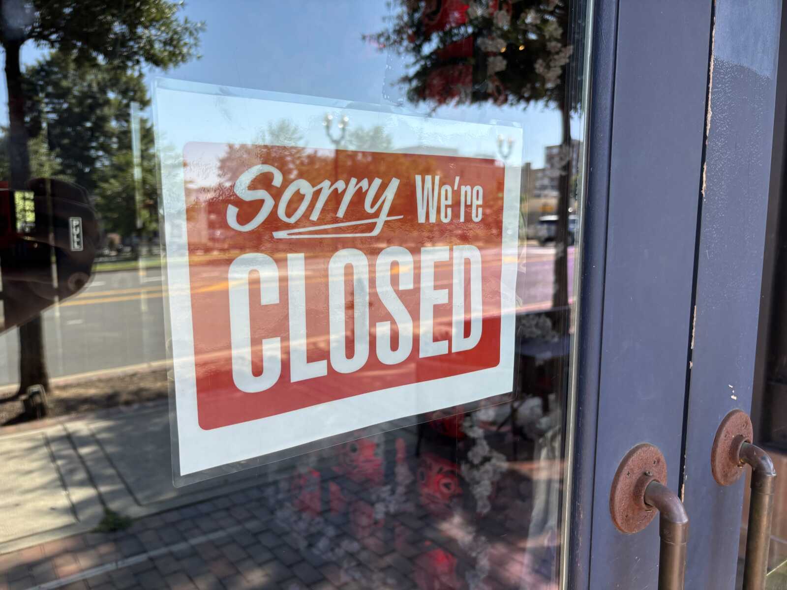 'Sorry We're Closed' sign on a glass door, indicating the shop is closed; reflections of the street visible in the glass.