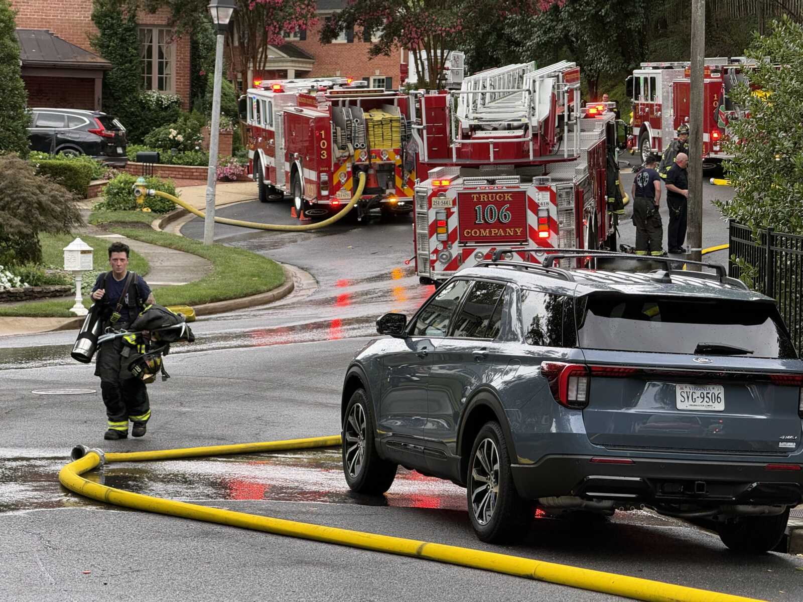 Firefighters and red fire trucks on a residential street, a large yellow hose running across the wet pavement toward a house beyond the SUV in the foreground.