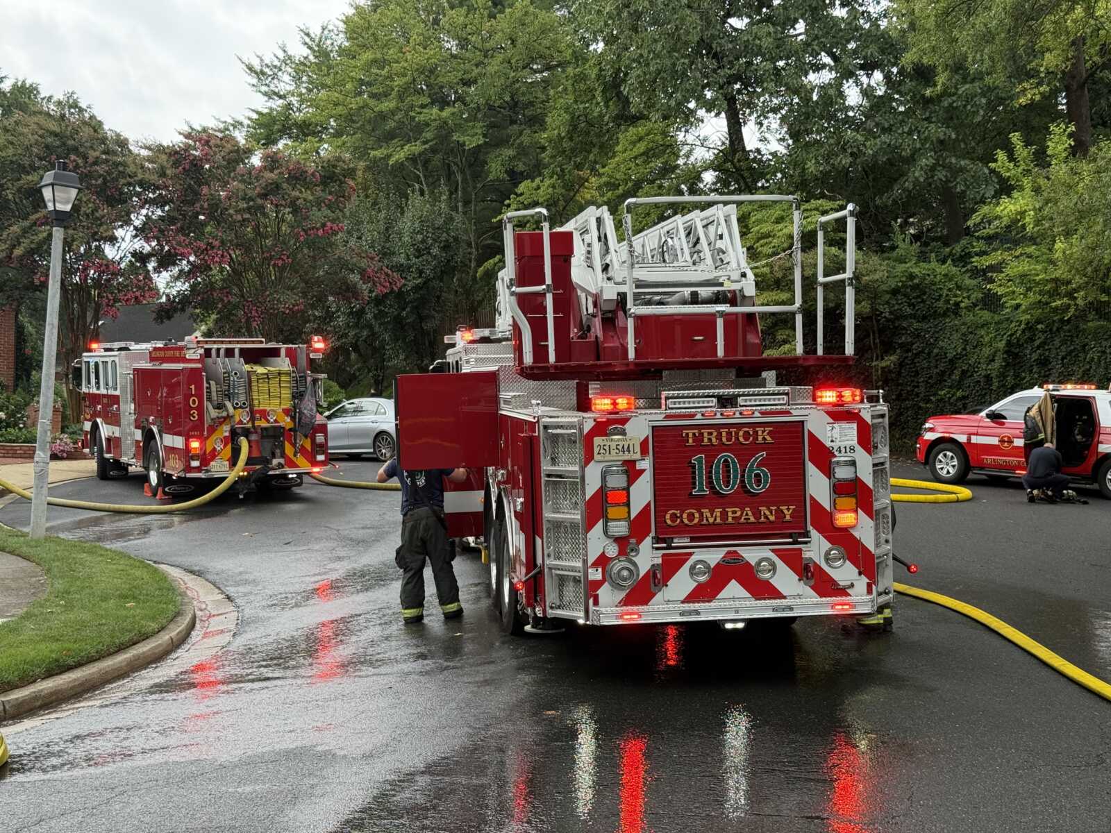 Fire trucks on a wet street with hoses deployed and firefighters at a brush-fire or structure fire scene.