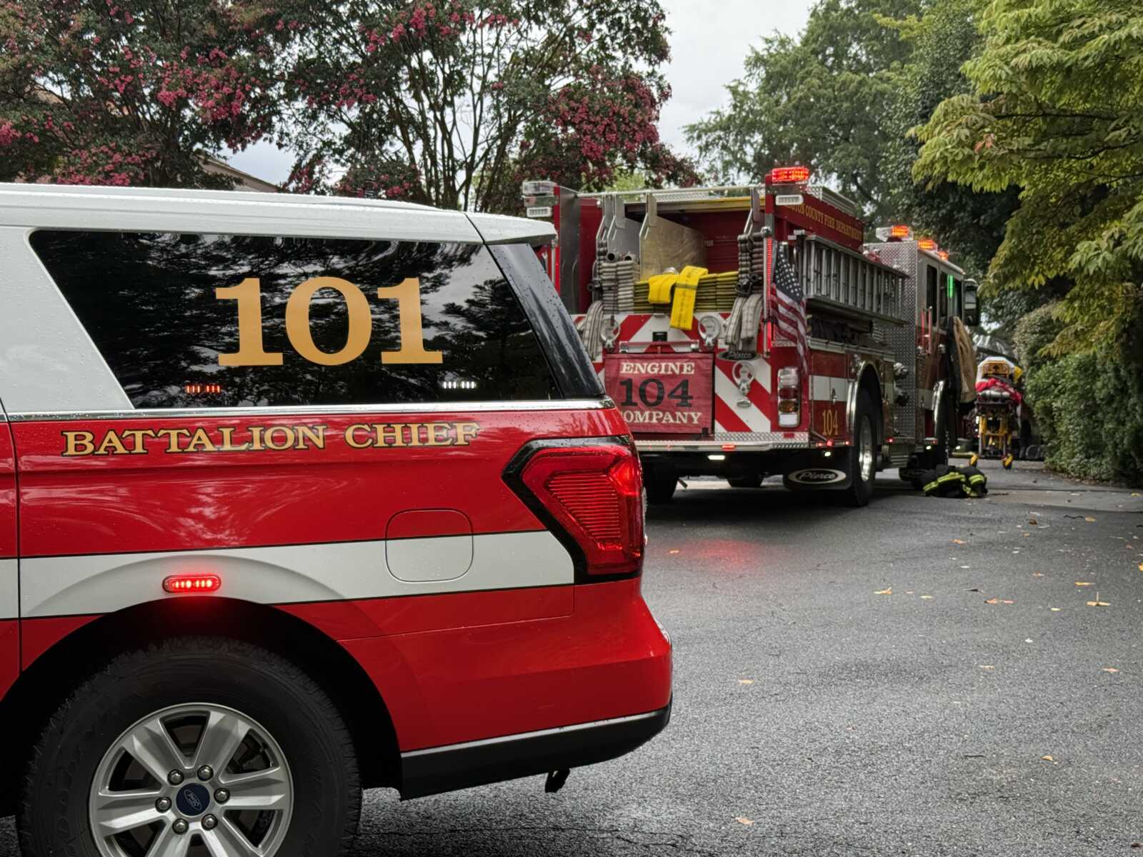 Fire department response scene with a red Battalion Chief SUV in the foreground and a large fire engine behind, parked on a residential street with equipment on the truck’s back deck.