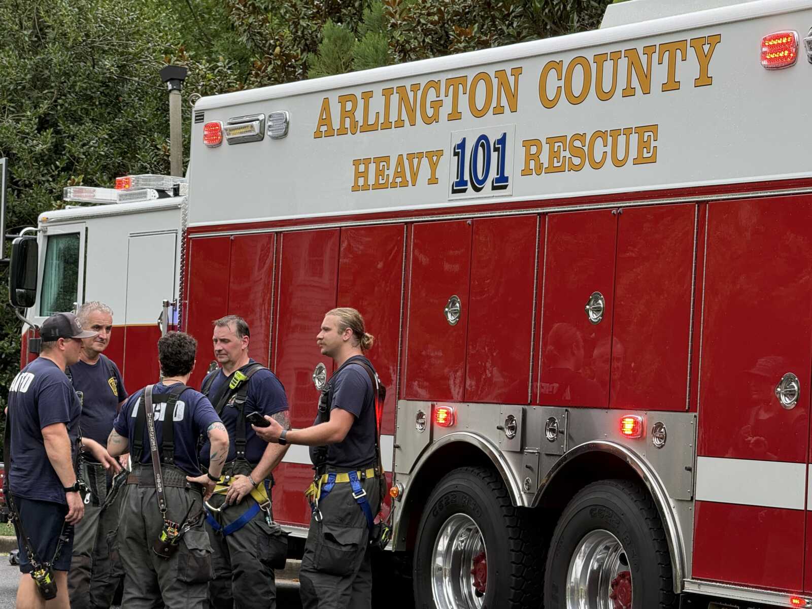 Group of firefighters in gear stand and talk beside a large red-and-white Arlington County Heavy 101 Rescue fire truck.
