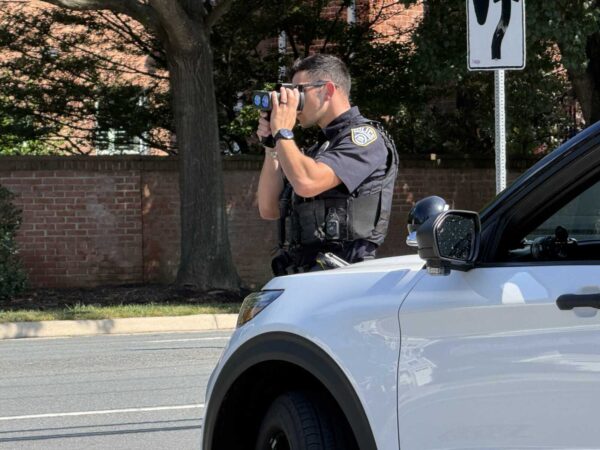 Police officer in tactical vest using binoculars from the side of a white patrol car on a street. (Informative)