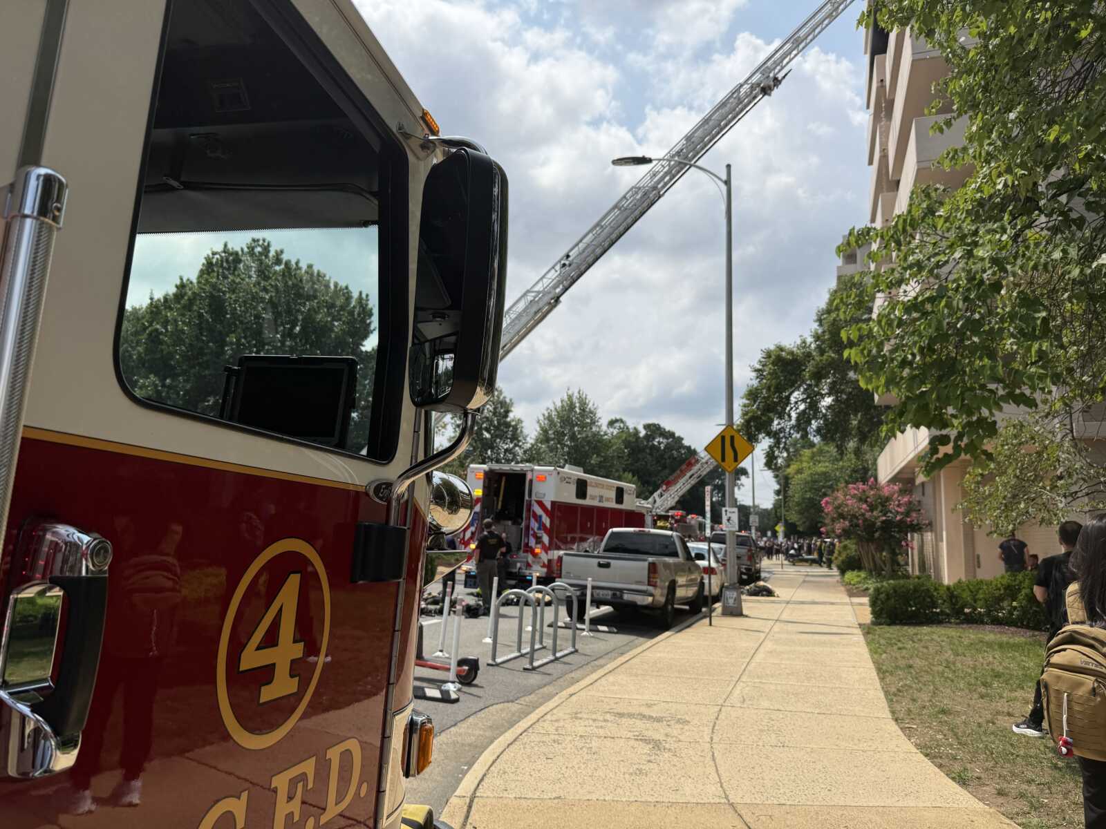 Fire trucks on a city street with a tall ladder extended toward a building, firefighters at the scene and onlookers nearby.