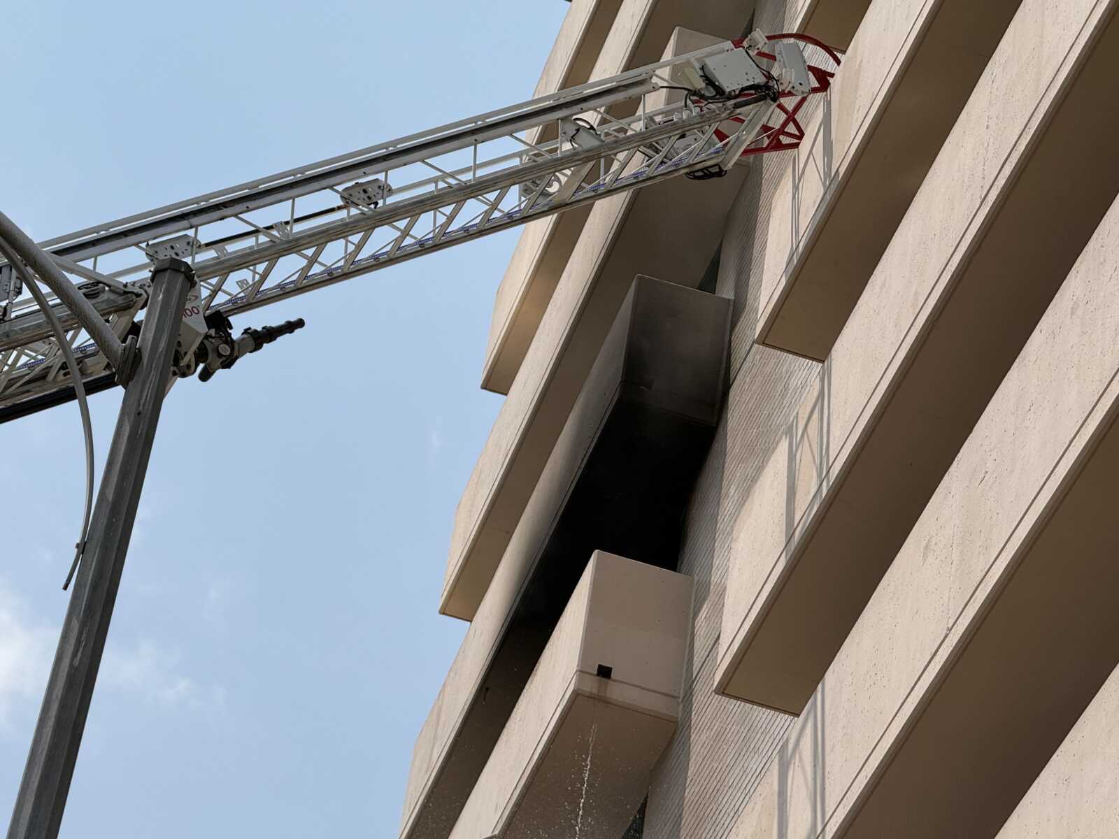 Aerial construction ladder/boom extending to the upper floors of a beige concrete building against a clear blue sky, showing metal lattice and cables