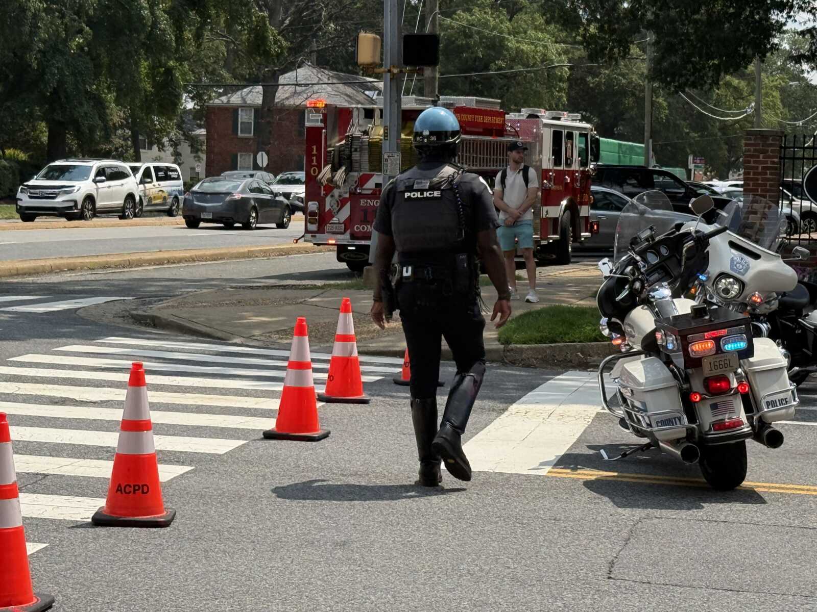 Police officer in a tactical vest walks through a crosswalk as orange cones line the street and emergency vehicles loom in the background.