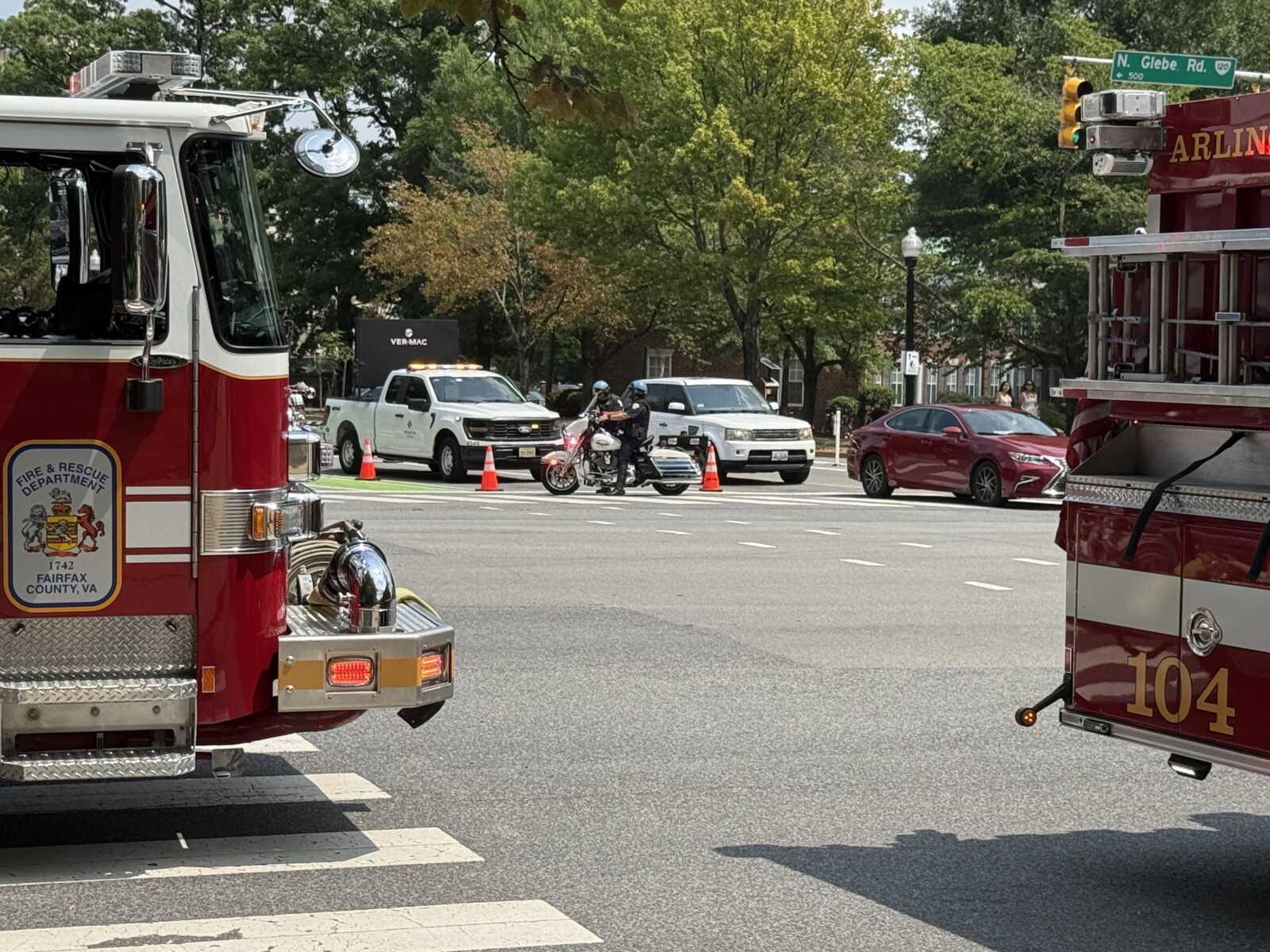 Fire trucks block a city intersection with orange cones, a police motorcycle, and other vehicles nearby; emergency scene on a sunny day in Virginia Weather: trees in background.