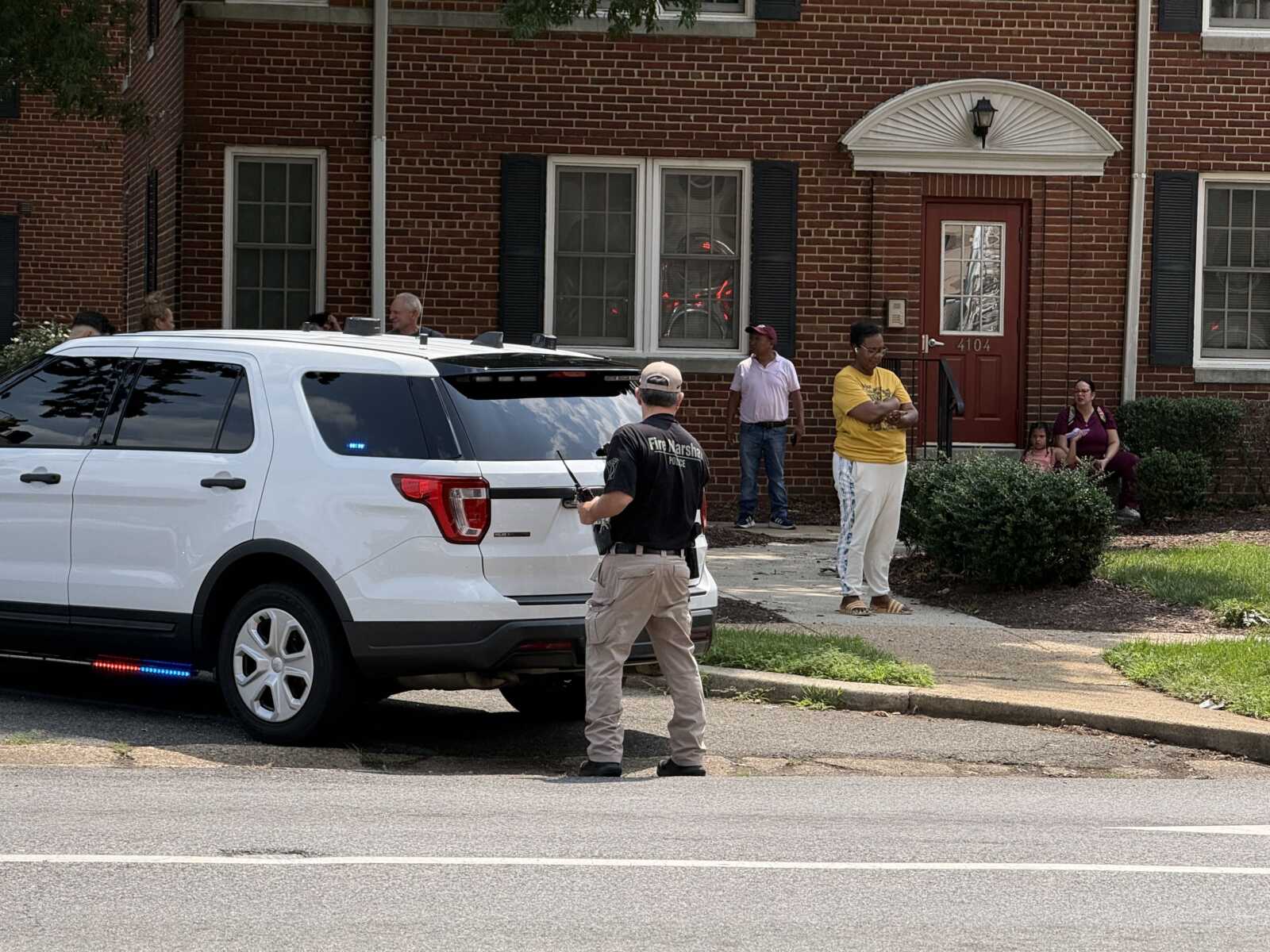 Fire department SUV with blue/red lights parked on a residential street as people stand outside an apartment building, likely assessing a situation.