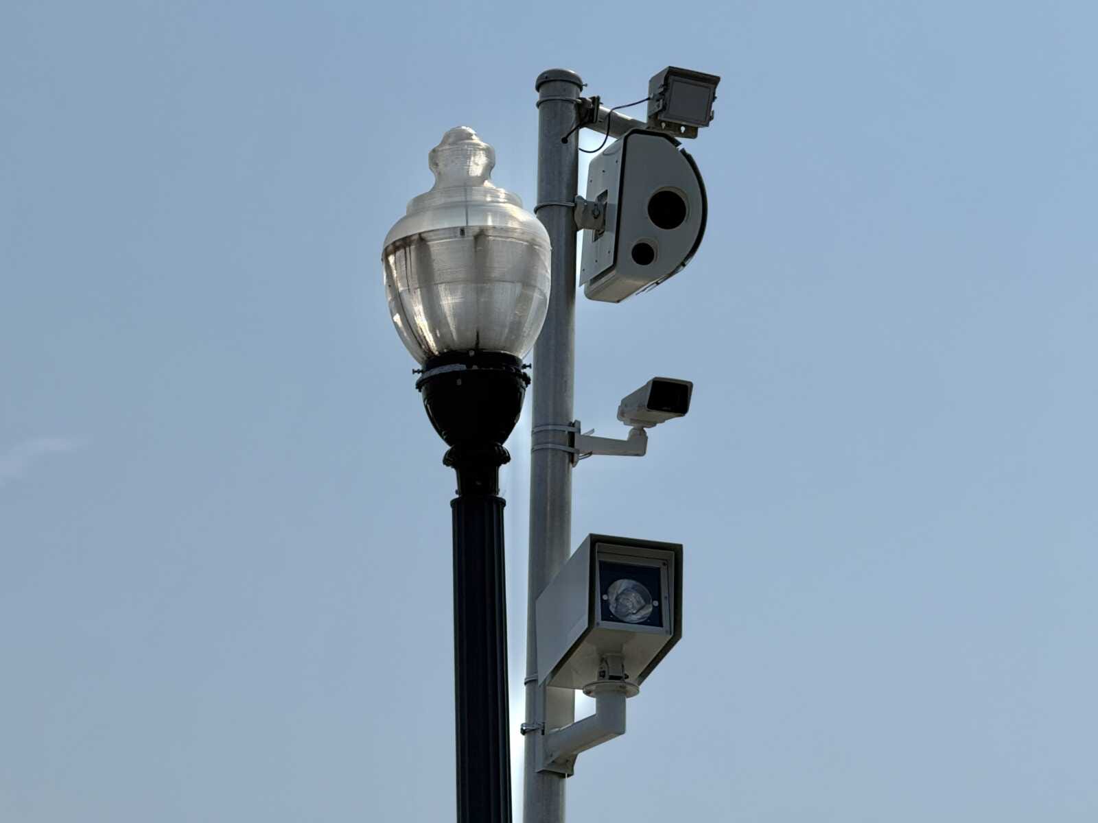 Red light camera along Langston Blvd (staff photo) | ARLnow Street lamp mounted on a metal pole with two security cameras against a clear blue sky.
