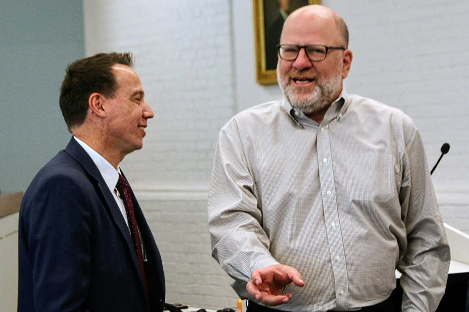 Two men conversing in a room: one in a suit, the other in a light button-down shirt, smiling as they talk near a podium microphone.