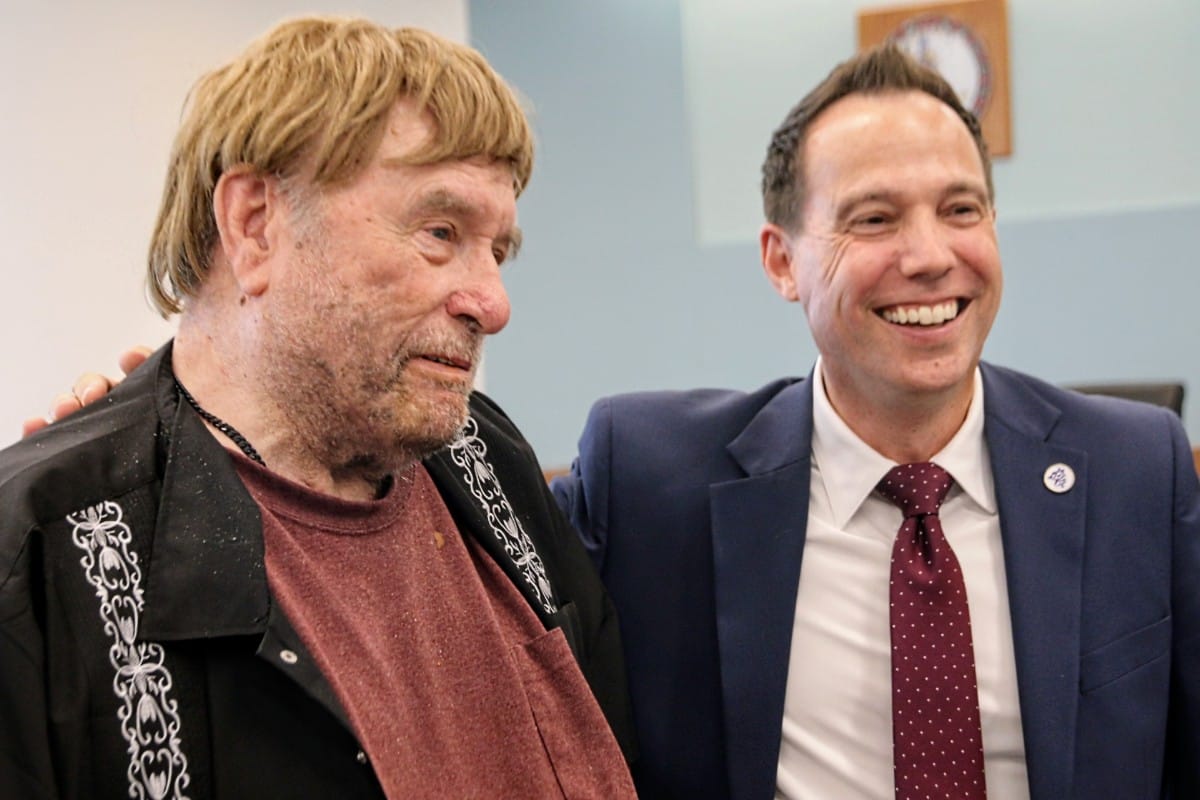 Two men smiling indoors, one older with blond hair and a decorative black jacket beside a younger man in a navy suit and maroon tie.