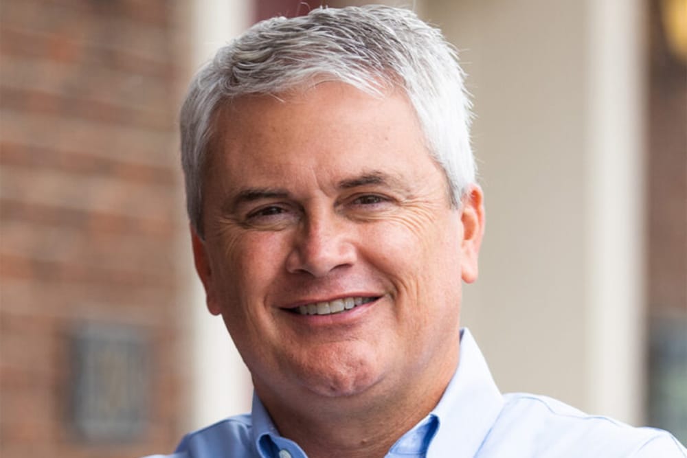 Smiling man with short gray hair wearing a light blue collared shirt outdoors.